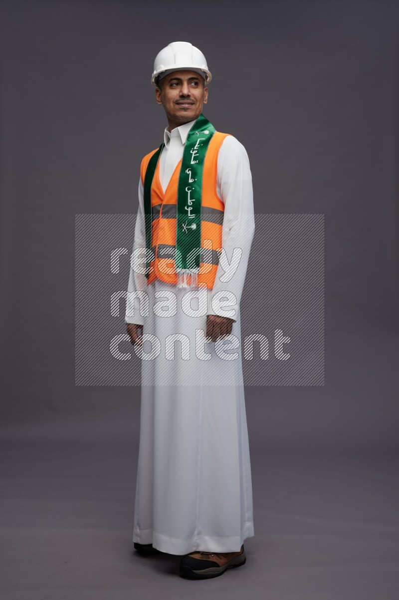 Saudi man wearing thob with engineer vest standing holding Saudi flag on gray background