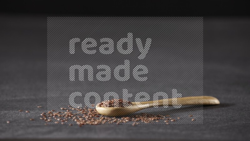 A wooden spoon full of flaxseeds and seeds spread beside it on a black flooring