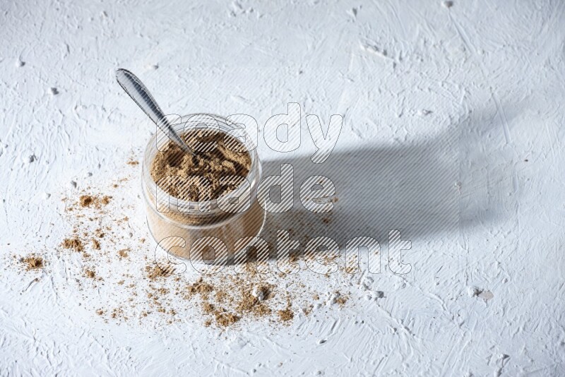 A glass jar and a metal spoon full of allspice powder on a textured white flooring