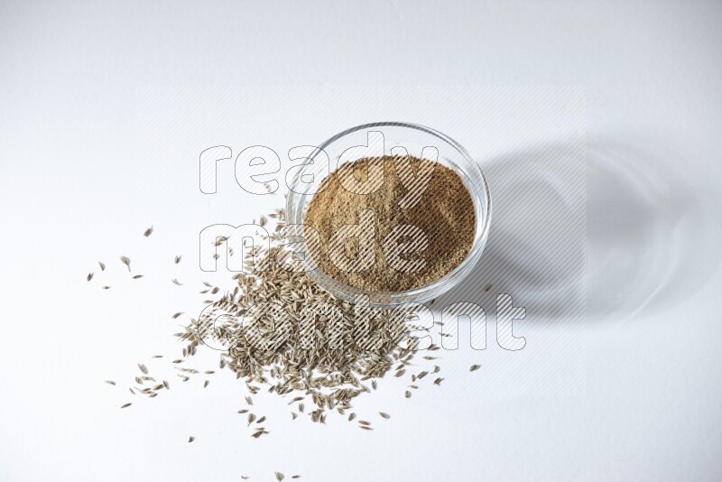 A glass bowl full of cumin powder with cumin seeds beneath it on white flooring