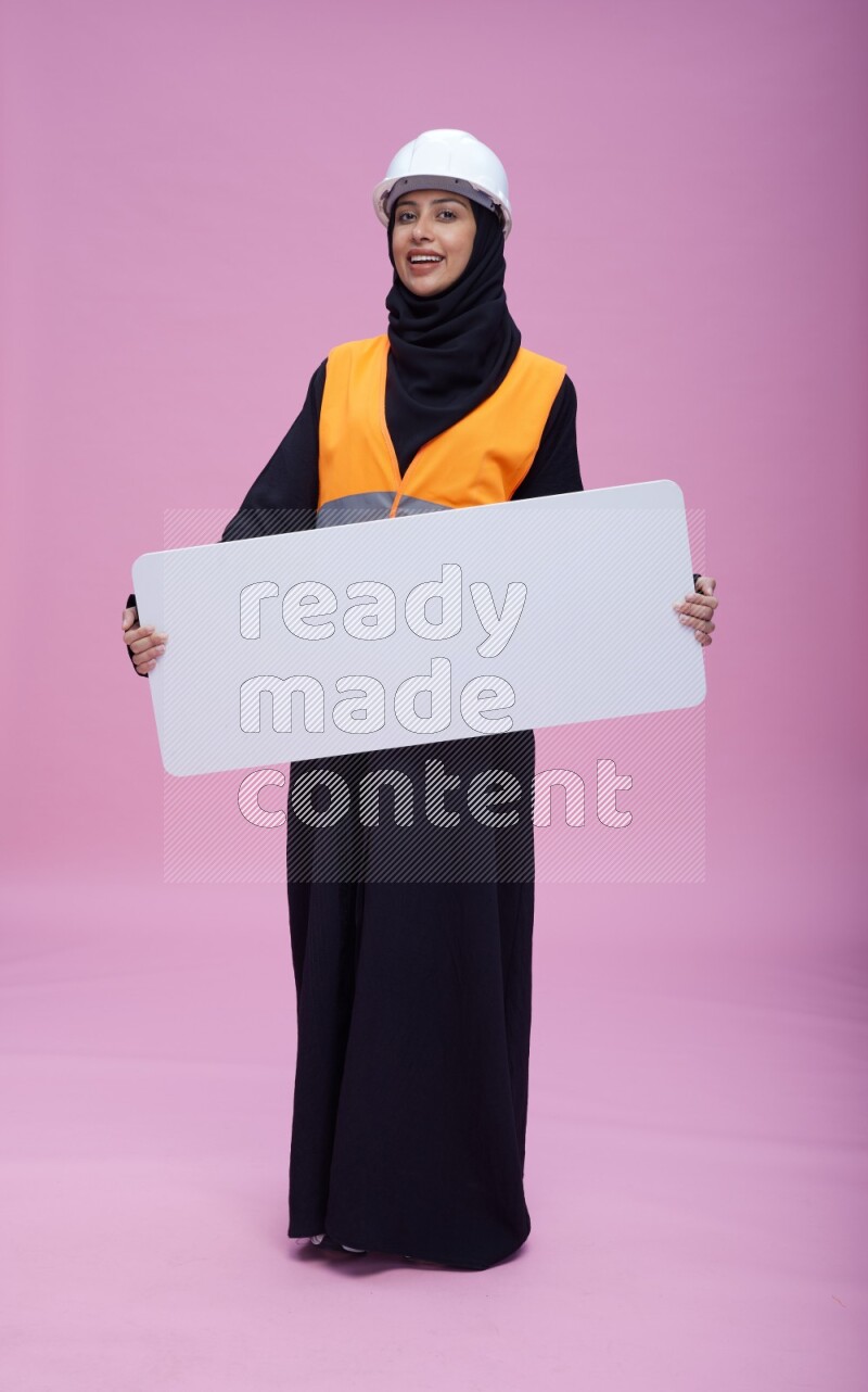 Saudi woman wearing Abaya with engineer vest and helmet standing holding board on pink background