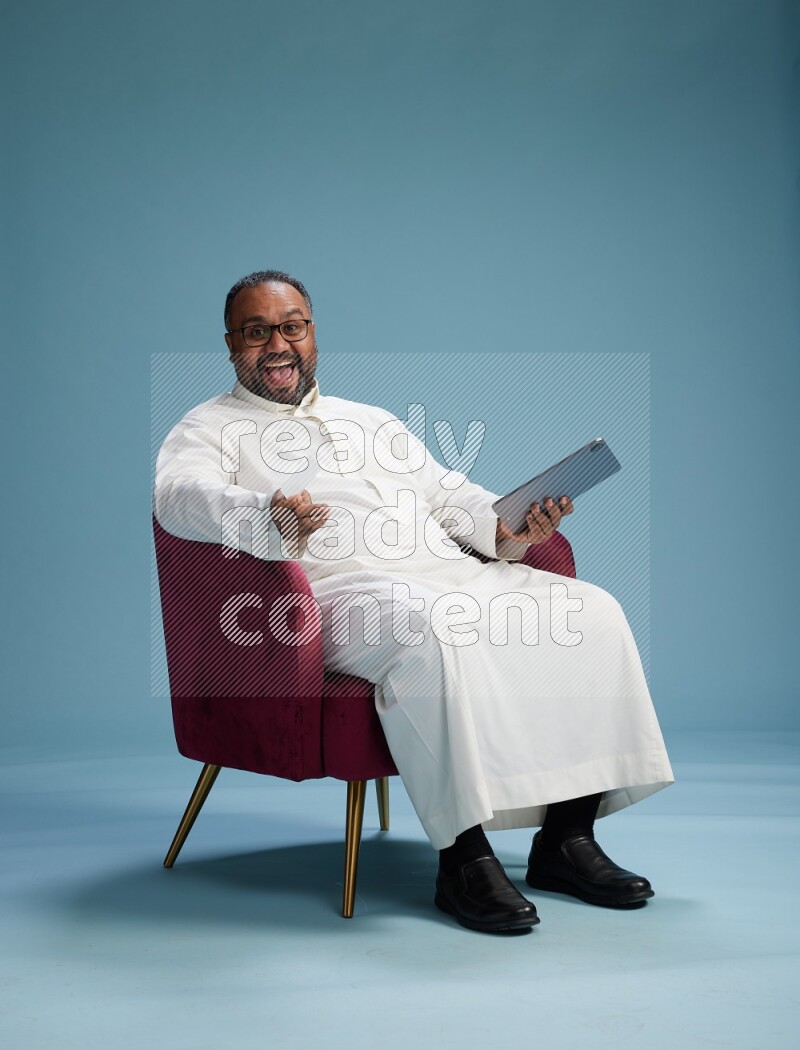 Saudi Man without shimag sitting on chair holding ATM card while working on tablet on blue background