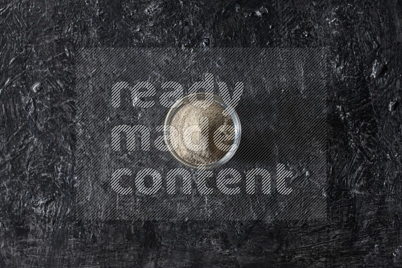 A glass bowl full of white pepper powder on textured black flooring