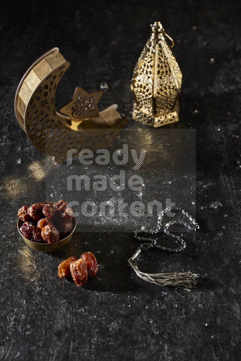 Dates in a metal bowl with prayer beads beside golden lanterns in a dark setup
