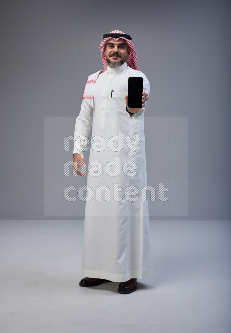 Saudi man Wearing Thob and red Shomag standing showing phone to camera on Gray background