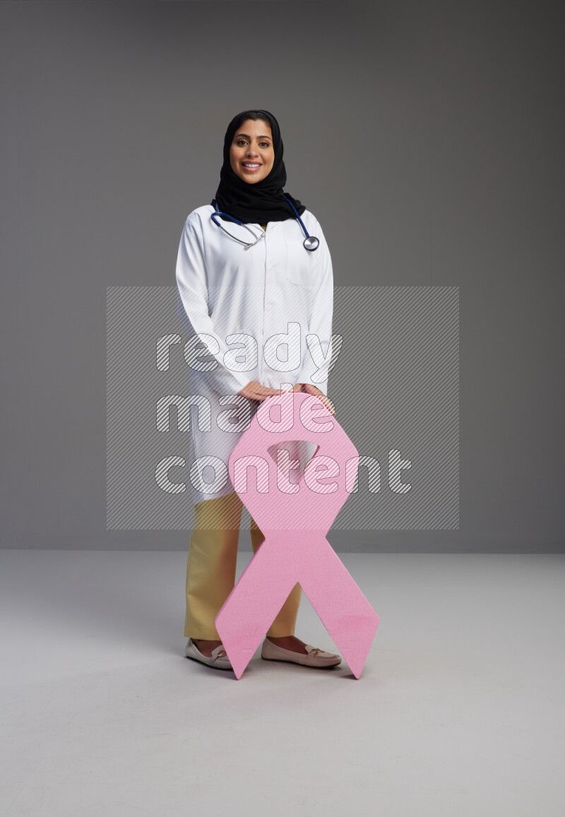 Saudi woman wearing lab coat with stethoscope standing holding awareness ribbon symbol standing on Gray background