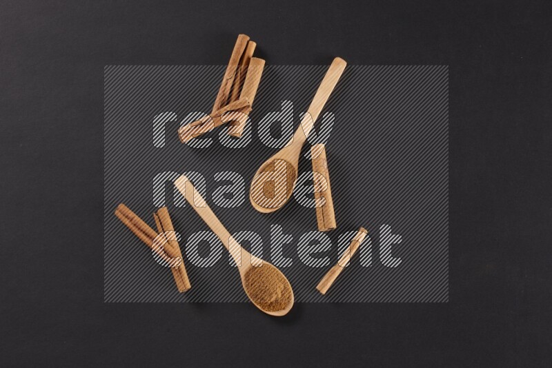 Cinnamon powder in two wooden spoons with cinnamon sticks on black background