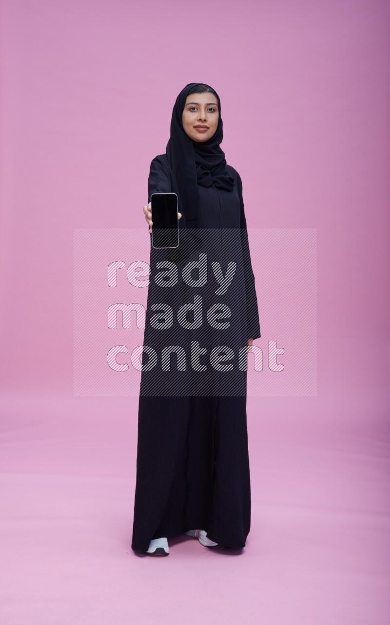 Saudi woman wearing Abaya standing showing phone to camera on pink background