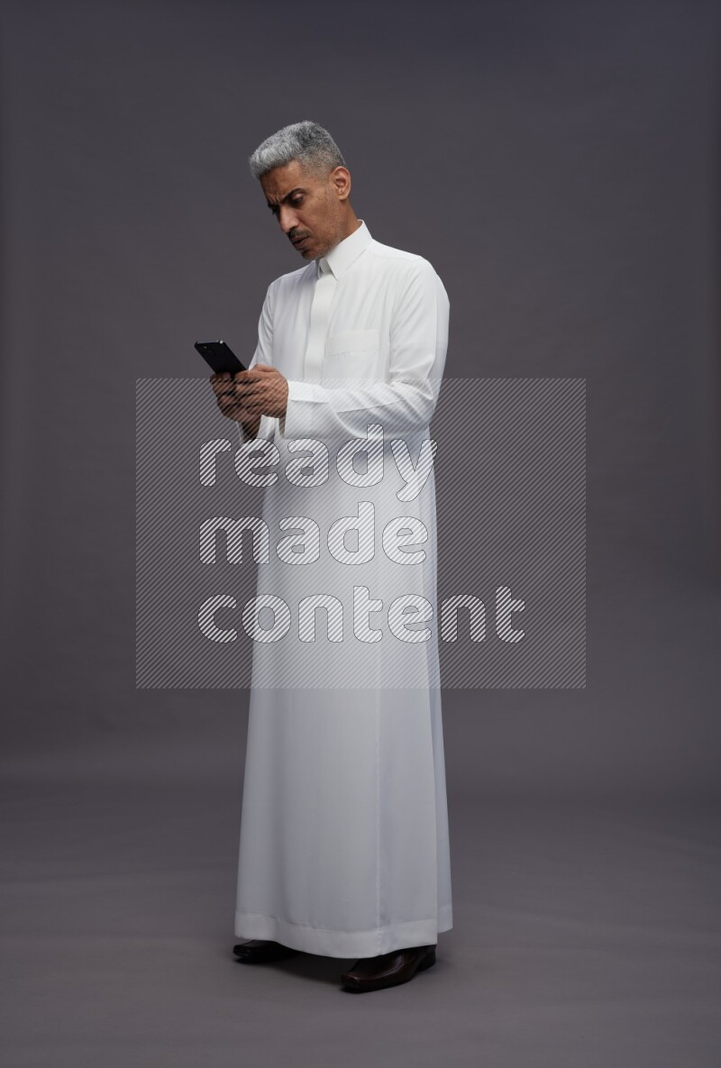 Saudi man wearing thob standing texting on phone on gray background