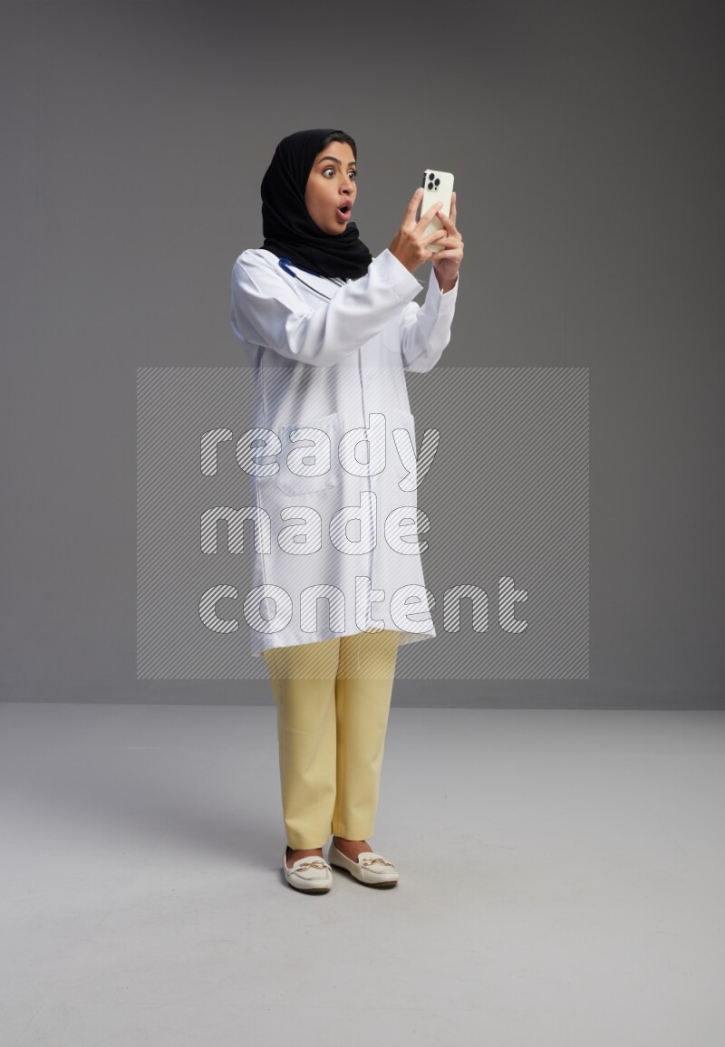 Saudi woman wearing lab coat with stethoscope standing texting on phone on Gray background