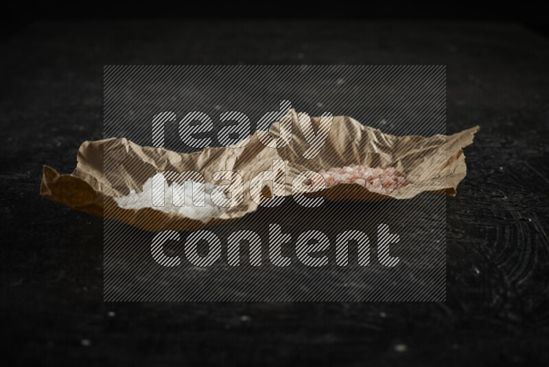 2 crumpled pieces of paper full of pink himalayan salt and white salt on black background