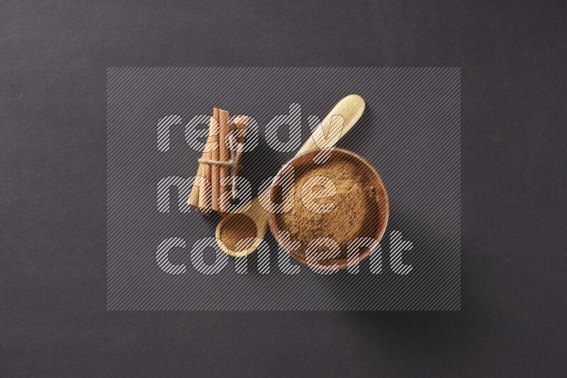 Cinnamon sticks stacked and bounded beside a wooden bowl full of cinnamon powder and a wooden spoon full of powder on black background