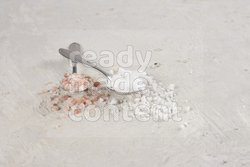 ملعقتان معدنيتان ممتلئتان بالملح الأبيض وملح الهيمالايا الوردي على خلفية بيضاء