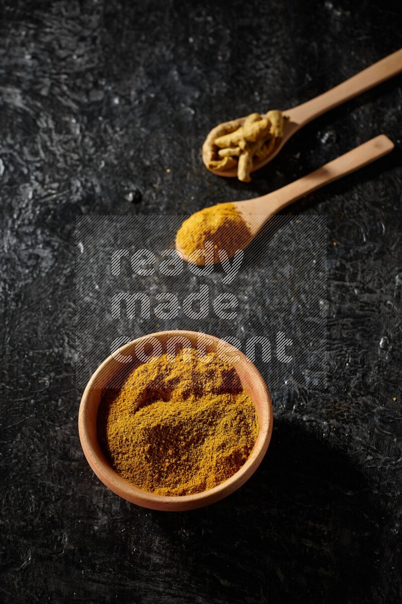 A wooden bowl full of turmeric powder and 2 wooden spoons full of dried turmeric finger and turmeric powder on textured black flooring