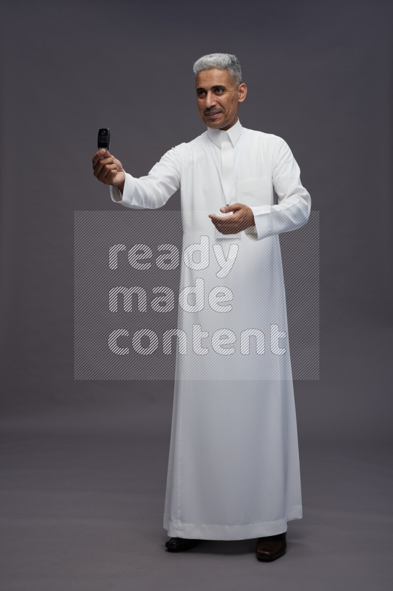 Saudi man wearing thob with neck strap employee badge standing holding key car on gray background