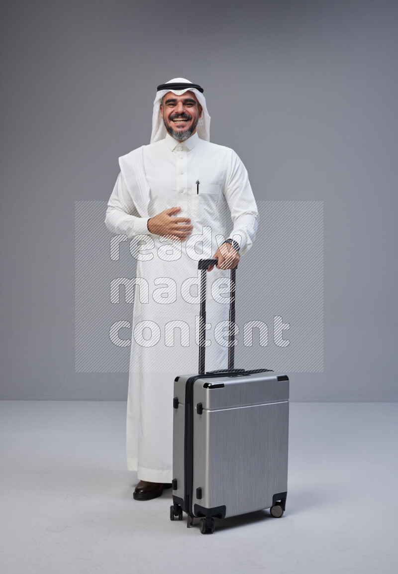Saudi man wearing Thob and white Shomag standing holding Travel bag on Gray background