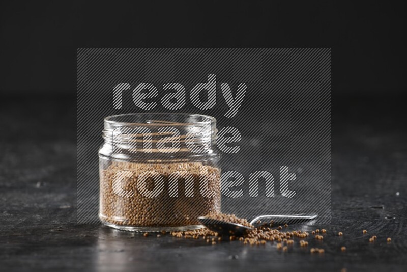 A glass jar and a metal spoon full of mustard seeds on a textured black flooring