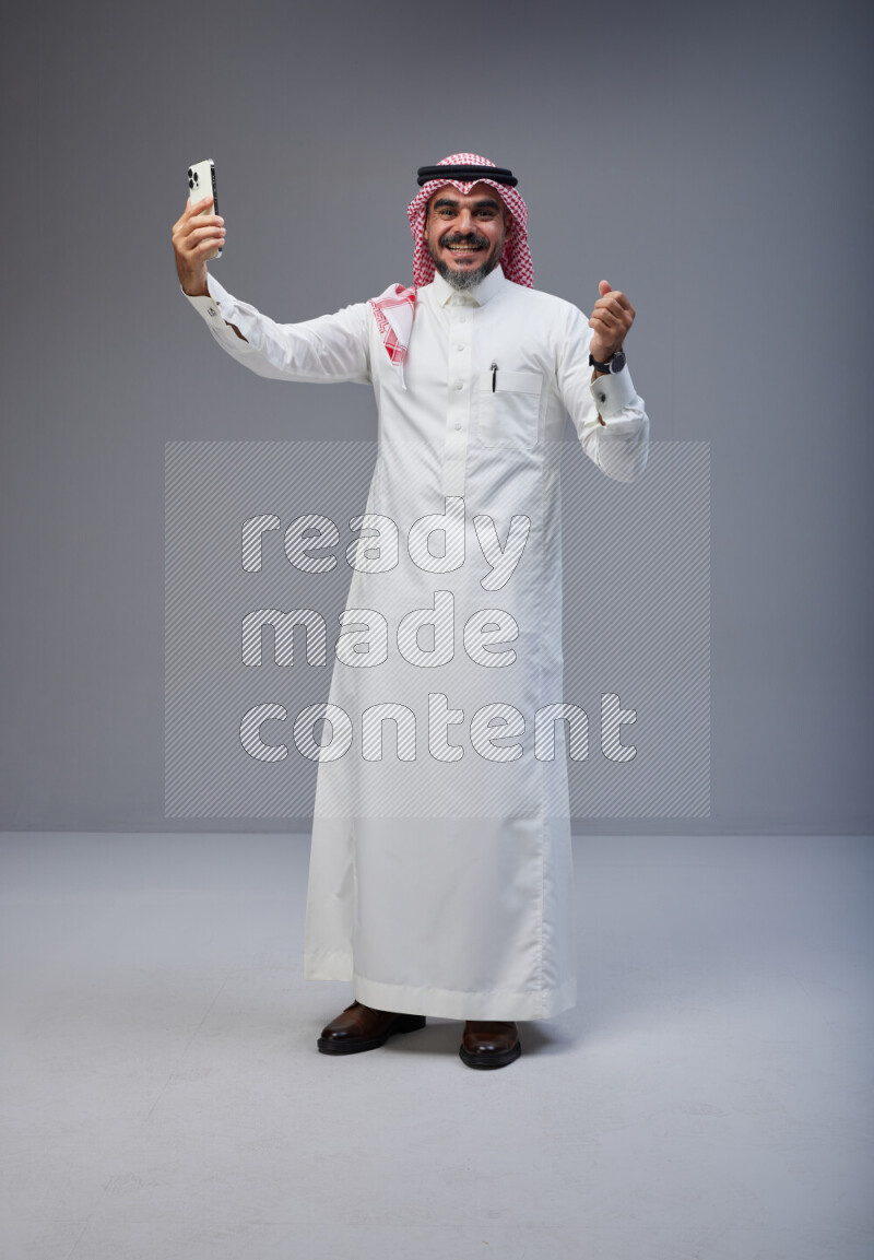 Saudi man Wearing Thob and red Shomag standing taking selfie on Gray background