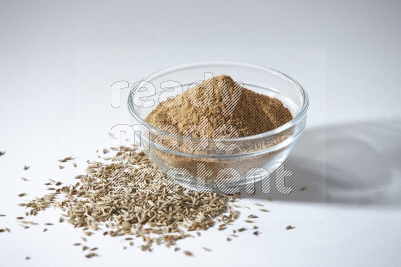 A glass bowl full of cumin powder with cumin seeds beneath it on white flooring