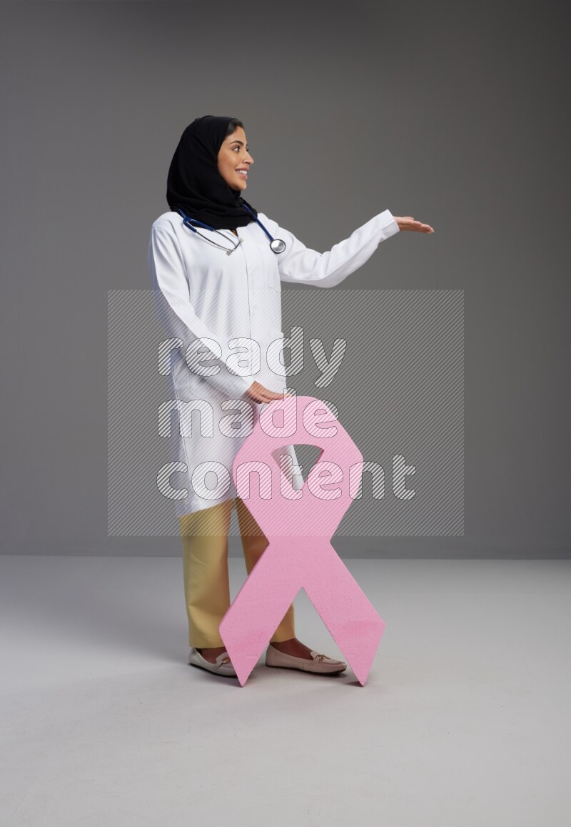 Saudi woman wearing lab coat with stethoscope standing holding awareness ribbon symbol standing on Gray background