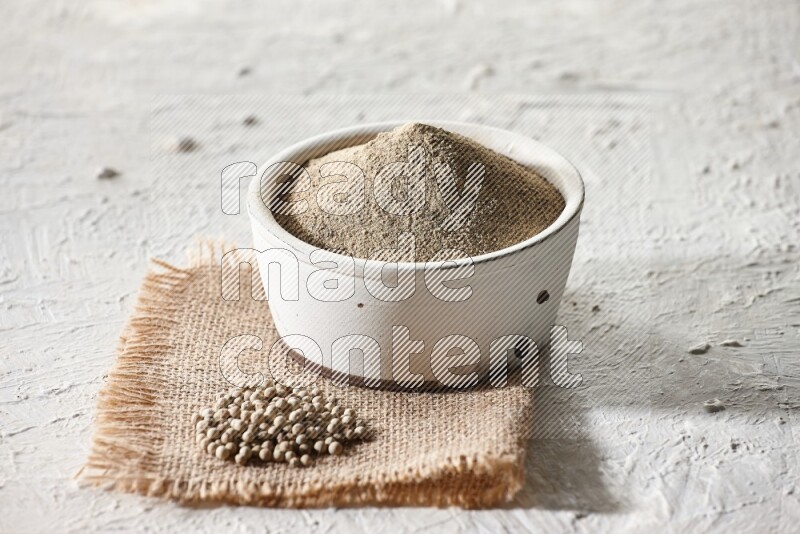 White pottery bowl full of white pepper powder set on a burlap piece of fabric with pepper beads and wooden pepper grinder on textured white flooring