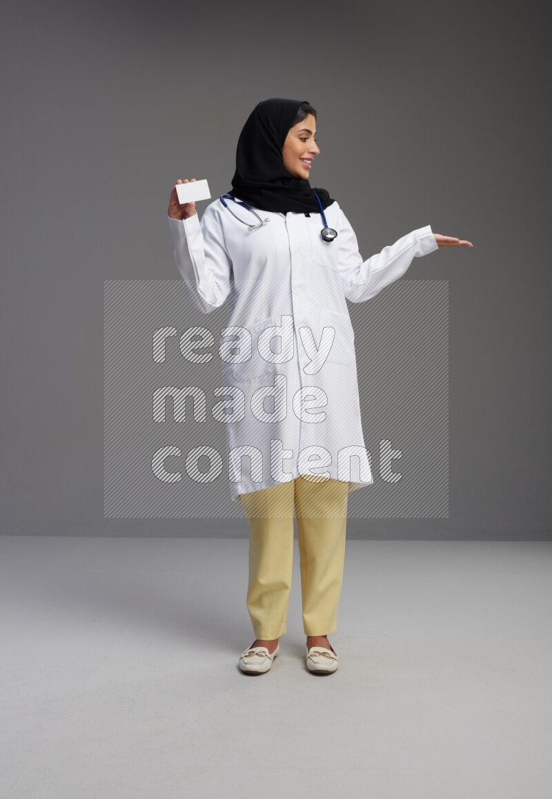 Saudi woman wearing lab coat with stethoscope standing holding ATM card on Gray background