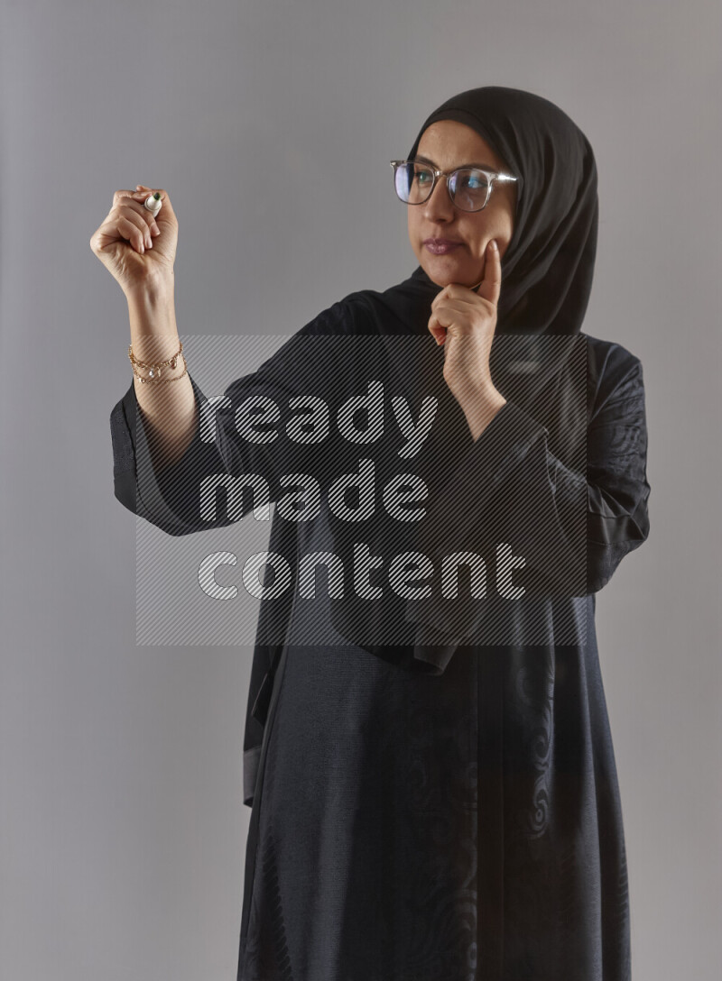 Woman in black abaya, hijab and glasses holding a marker pen to write on a transparent board with different reactions and expressions on grey background