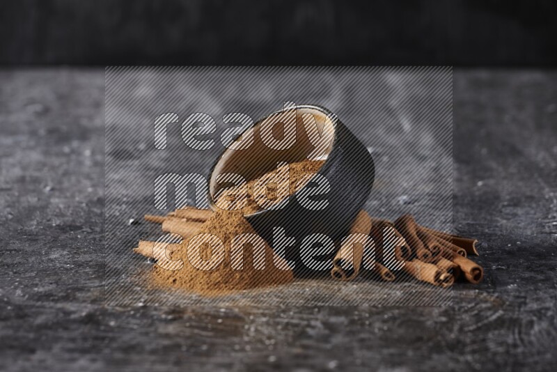 Black pottery bowl over filled with cinnamon powder and cinnamon sticks around the bowl on a textured black background