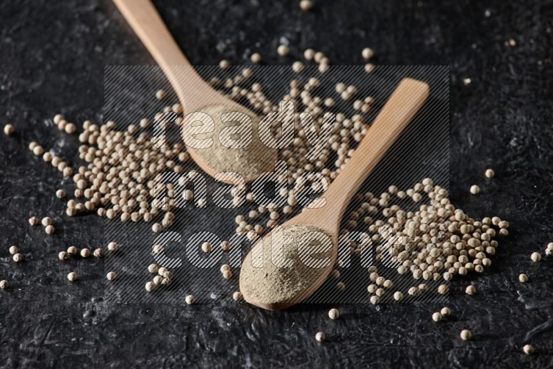 2 wooden spoons full of white pepper powder with white pepper beads on textured black flooring