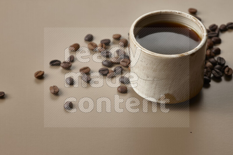 A beige pottery cup of coffee surrounded by roasted coffee beans on beige background