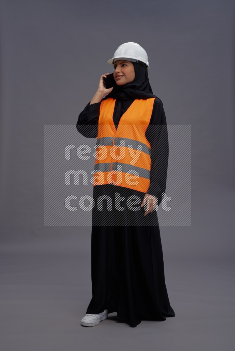 Saudi woman wearing Abaya with engineer vest standing talking on phone on gray background