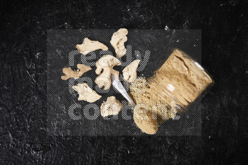 A glass jar full of ground ginger powder flipped with some spilling powder on black background