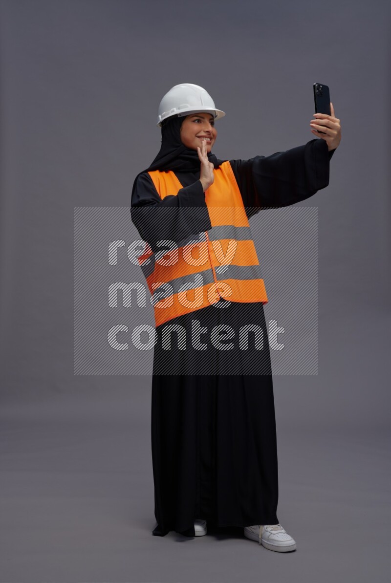 Saudi woman wearing Abaya with engineer vest standing taking selfie on gray background