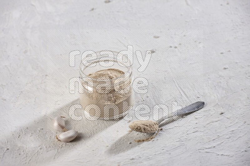 A glass jar full of garlic powder with a metal spoon full of the powder on a textured white flooring