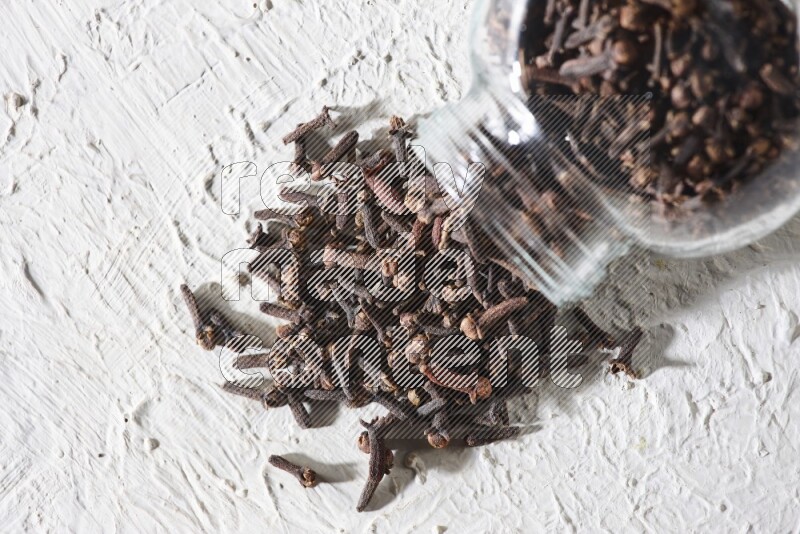 A flipped glass spice jar full of cloves and cloves came out of it on textured white flooring