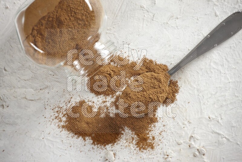 Flipped herbs glass jar full of cinnamon powder with a metal spoon full of powder on a textured white background