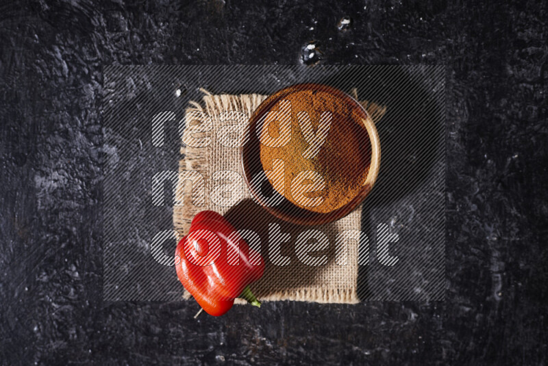 A wooden bowl full of ground paprika powder with a red bell pepper on a burlap fabric on black background