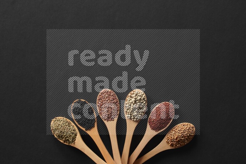 wooden spoons filled with white peppers, mustard seeds, black seeds, garden cress, cumin and flax on black flooring