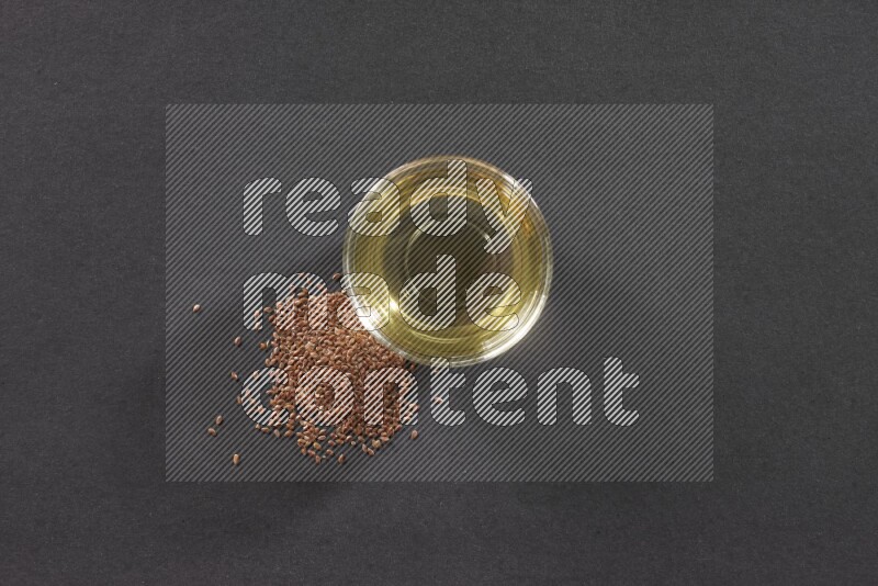 A glass bowl full of flaxseeds oil with some of flaxseeds beside it on a black flooring