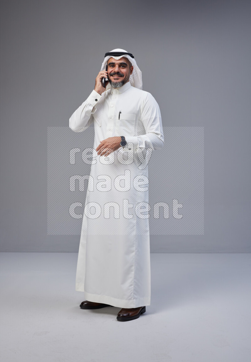 Saudi man Wearing Thob and white Shomag standing talking on phone on Gray background