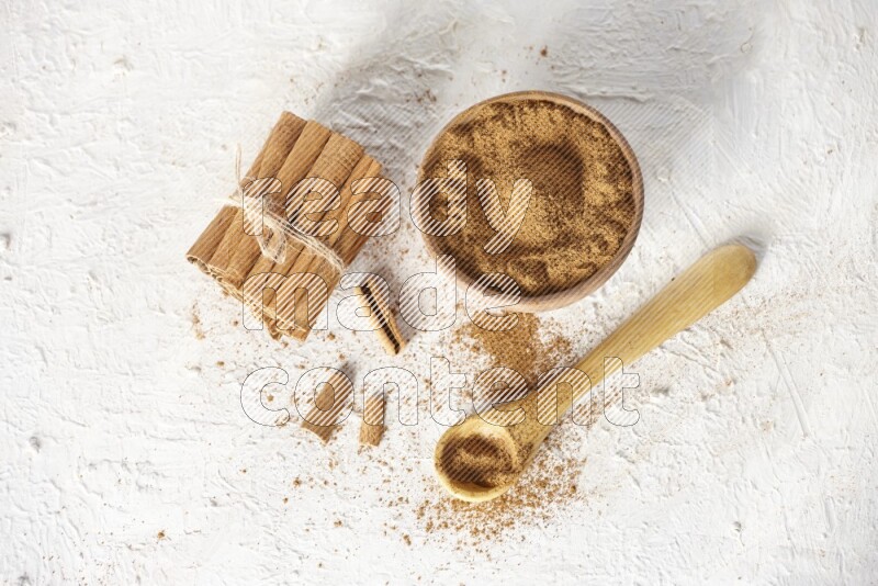 Cinnamon sticks stacked and bounded beside a wooden bowl full of cinnamon powder and a wooden spoon full of powder on white background
