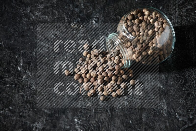A flipped glass spice jar full of allspice whole balls and the balls spilled out of it on a textured black flooring