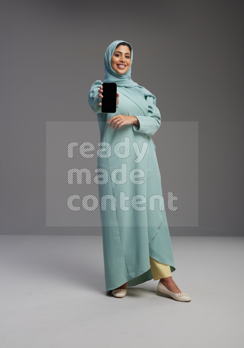 Saudi Woman wearing Abaya standing showing phone to camera on Gray background