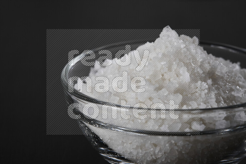 A glass bowl full of coarse sea salt crystals on black background