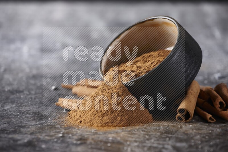 Black pottery bowl over filled with cinnamon powder and cinnamon sticks around the bowl on a textured black background