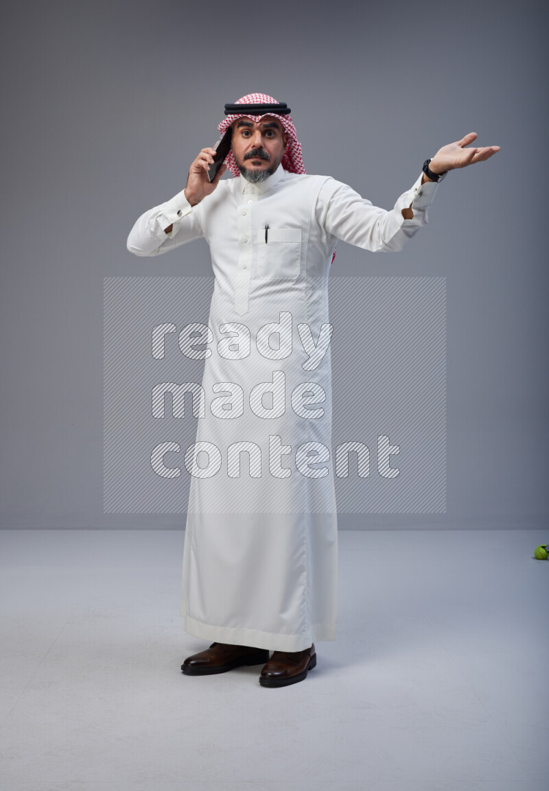 Saudi man Wearing Thob and red Shomag standing talking on phone on Gray background