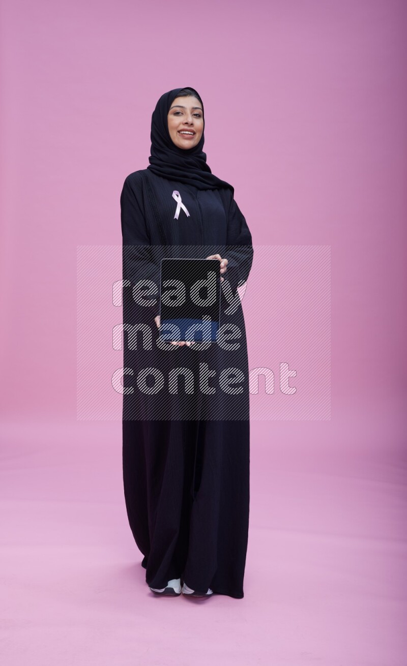Saudi woman wearing pink ribbon on Abaya standing showing a tablet to camera on pink background