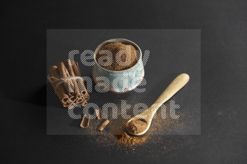 Ceramic bowl full of cinnamon powder and a wooden spoon full of powder with cinnamon sticks stacked and bounded on black background
