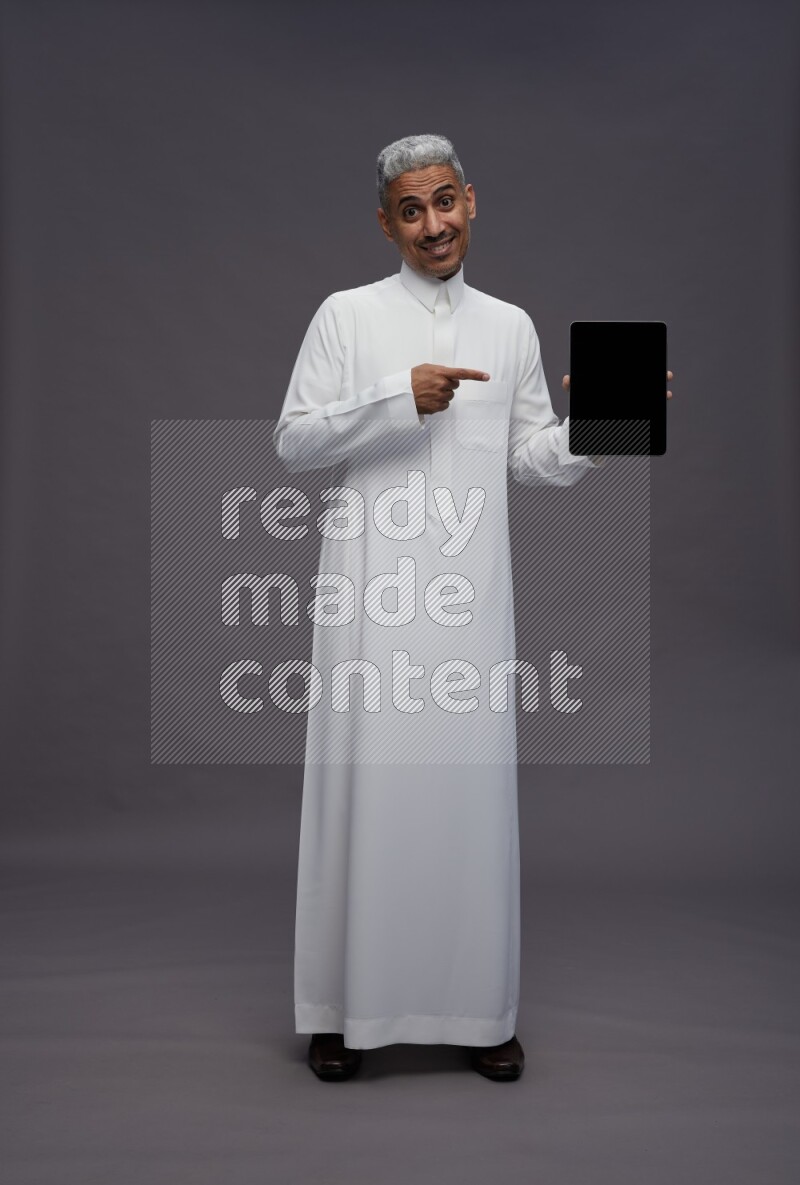 Saudi man wearing thob standing showing tablet to camera on gray background