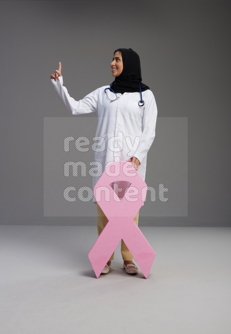 Saudi woman wearing lab coat with stethoscope standing holding awareness ribbon symbol standing on Gray background